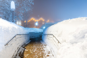A paved path with stainless steel handrails cleared of snow among deep snowdrifts in an alpine village illuminated by artificial light in the late evening
