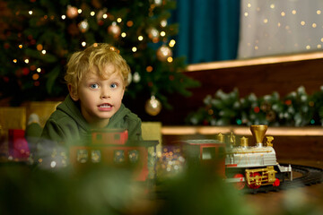 Happy curly-haired boy is playing with a magic toy train at the Christmas tree. The concept of...