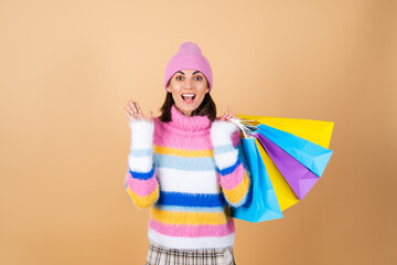 Young woman on a beige background in a bright multi-colored cozy knitted sweater, a plaid skirt holds shopping packages shouts in surprise excitedly