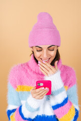 Young woman on a beige background in a knitted sweater and a hat with a mobile phone laughing with surprise and fun, reading messages, jokes, congratulations