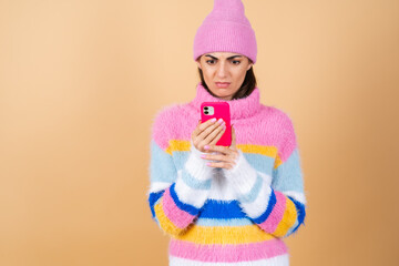 Young woman on a beige background in a knitted sweater and a hat with a mobile phone looks at the screen with disgust displeasure