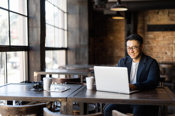 Asian businessman typing on laptop during work in cafe. Concept of remote and freelance work. Smiling adult successful man wearing suit and glasses sitting at wooden desk. Sunny day - obrazy, fototapety, plakaty