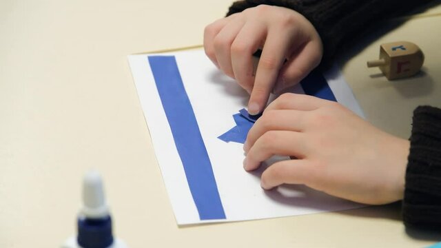 Little Child Sitting at the Desk and makes the Israel flag with paper and glue. Glues pieces of blue paper onto cardboard with hands and make Israel symbol. Jewish religion symbol. 