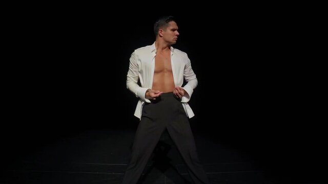 Young male ballroom dancer with bare breast,strong pectoral muscles,abs,in open white shirt,black trousers on dark backdrop,expressing passion.Solo dance and search for dance partner concept,midpoint.