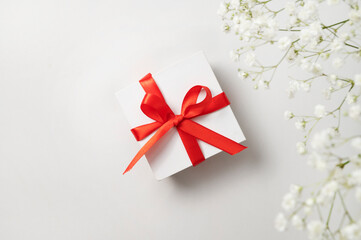 a white gift box with a red ribbon on a white  background with a bouquet of  gypsophila. The concept of a holiday or gift card. Top  view and copy space