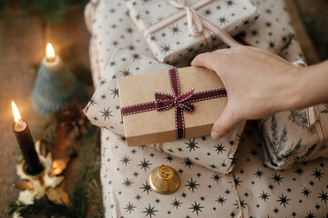 Hand opening stylish christmas gift wrapped in paper on rustic wooden table with candles, bells, fir branches. Stylish eco xmas presents, atmospheric moody image. Happy Holidays!
