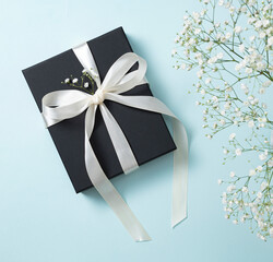 a black gift box with a white ribbon on a blue background with a bouquet of  gypsophila. The concept of a holiday or gift card. Top  view and copy space