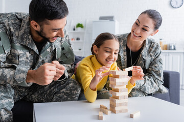 Happy parents in military uniform showing yes gesture near kid playing wood blocks game at home