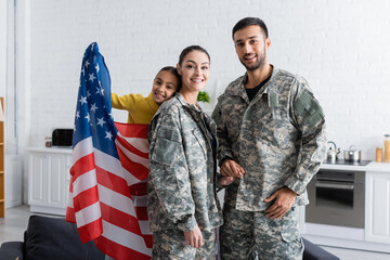 Parents in military uniform holding hands near preteen kid with american flag at home