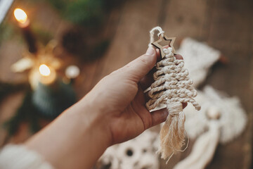 Hand holding stylish christmas tree macrame ornament on background of candles, fir branches on rustic wooden table. Handmade boho ornament for xmas holiday. Scandinavian decor - obrazy, fototapety, plakaty