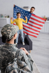 Woman in military uniform with backpack near blurred family holding american flag in kitchen