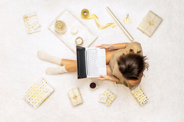 Christmas. Online shopping. Woman in sweater using laptop for making order, buying, sitting on the white carpet among the many wrapped gifts in white gold wrapping paper. Top view. Concept