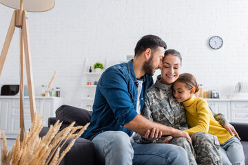 Smiling soldier in uniform sitting near child and husband on couch at home