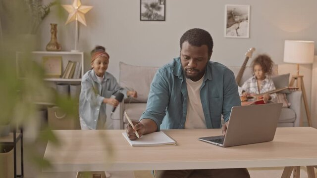 Medium slowmo shot of African-American man trying to work on laptop from home with two little daughters singing in microphone and playing guitar in background