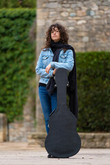  Young woman with curly hair and glasses posing with a guitar case in the street