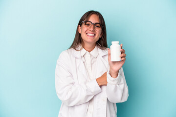 Young pharmacist woman holding pills isolated on blue background laughing and having fun.