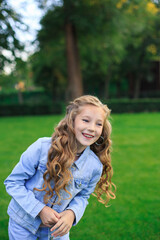 Obraz premium Blond-haired, blue-eyed girl in a blue suit against a green park background. A joyful portrait of a girl in an open space.
