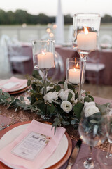 Lit candles in transparent glass jars on a wooden table A glass vase of water with burning candle inside decorated by green on the table Wedding table set decor 