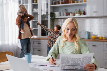 smiling woman with newspaper writing in notebook while husband playing with kids on blurred...
