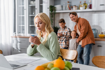 busy woman with cup of tea working at laptop near blurred fruits and cheerful family in kitchen