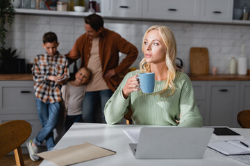 thoughtful woman with cup of tea sitting near laptop, documents, and blurred husband with kids in...