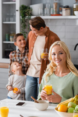 cheerful woman looking at camera near blurred husband and sons in kitchen