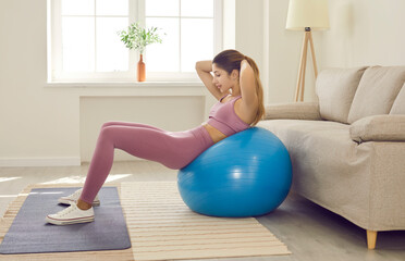 Woman having fitness workout at home. Young girl using exercise ball for abs training. Side view of happy lady holding hands behind head and doing sit ups on stability ball near sofa in living room