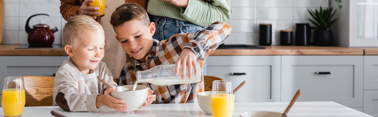 boy pouring milk during breakfast with brother near parents, banner