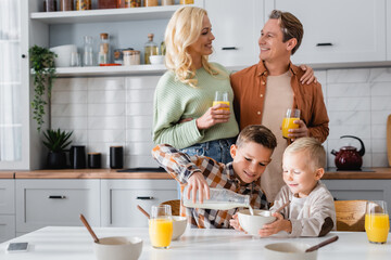 cheerful couple with glasses of orange juice near kids having breakfast in kitchen