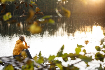 Woman in a yellow jacket relaxing on a wooden pier on a lake with a glass of rose wine watching fall sunset alone.