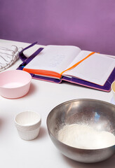 In the foreground is a bowl of flour and cupcake tins. In the background is an open notepad. Selective focus.