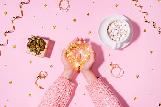 Beautiful holiday concept. Hands holding string garland lights, on the background of wrapped gift, a cup of hot chocolate or cocoa with marshmallows, golden tinsel isolated on pink background.