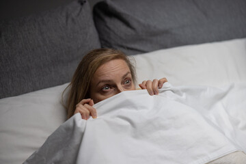 Scared caucasian woman lying on comfy bed and hiding under white soft blanket. Mature lady with blond hair suffering from insomnia because of nightmares.