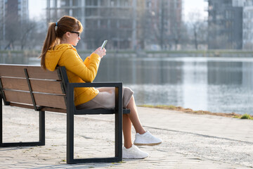 Young pretty woman sitting on a park bench browsing her smartphone outdoors on warm autumn day.