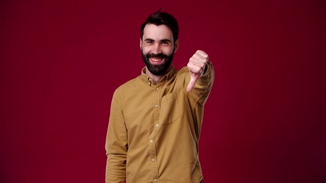 the guy shows a dislike at the camera, he laughs and is happy about it, he is wearing a brown shirt, black-haired, bearded, unshaven, background red
