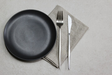 Black empty plate with knife and fork with gray textile napkin on grey stone background. Table setting. Top view. 