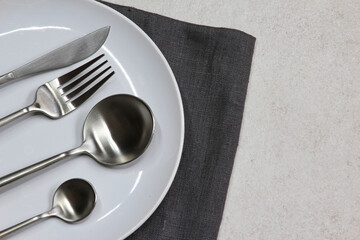 White empty plate with knife, spoons and fork dark textile napkin on grey stone background. Table...
