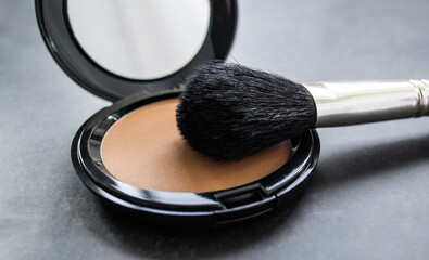 Close-up a black makeup brush next to an open box with a mirror and powder puff and pink powder....