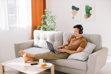 Relaxed young guy in wireless headphones lying on sofa in minimalistic living room and using laptop while working online