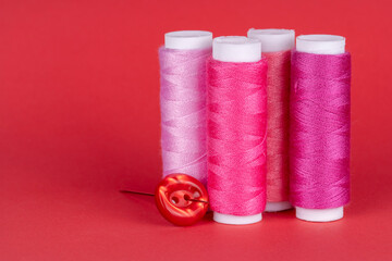 Four bobbins of sewing thread of different shades of pink with a red button and a needle isolated on red