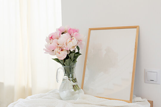 Vertical frame mockup on a wooden table in the kitchen. Glass vase with a bouquet of pink peonies. Scandinavian style interior.
