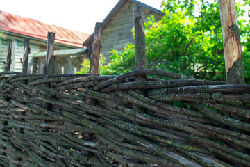 Fototapeta na wymiar An old wicker fence made of branches against the background of old wooden houses and green bushes. Daylight. Close-up.