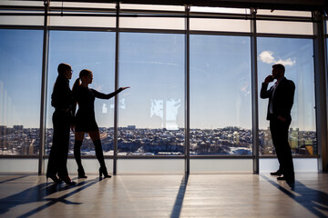 Two women and a man in the office enjoy the view of the city from the window of a skyscraper and talk while standing by the large window in the office