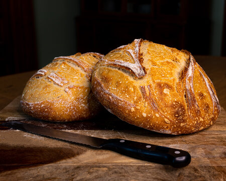 Two Whole Round Loaves of Crusty Artisan Bread on a Wooden Board in a Dark Room