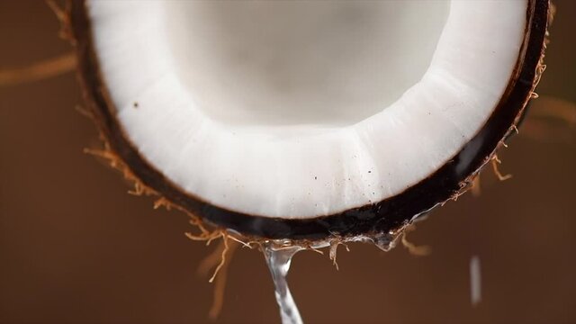 Coconut pouring water, dripping coconut milk, drops of coco nuts oil over brown background. Tropical Coco nut closeup. Healthy Food, skin care concept. Vegan food. Slow motion. 