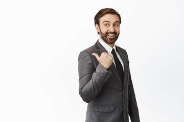 Enthusiastic businessman in suit pointing at product, empty space for company banner, standing over white background, smiling