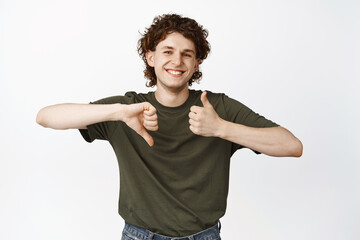 Smiling curly-haired young man showing thumbs up and thumbs down, dislike like gesture, leave...
