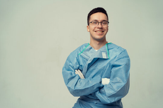 A Male Doctor In A Surgeon's Uniform. Posing For The Camera In The Studio On A Gray Background. Portrait Of A Smart Man With Glasses.