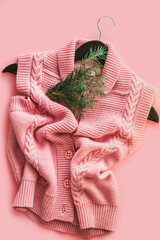 Knitted pink vest on a black wooden hanger on a pink background. Spruce twig and cozy knitted clothes of the autumn-winter period. Advertising banner for a clothing boutique.