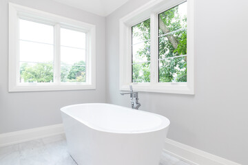 Fototapeta na wymiar A beautiful white standalone bath tub with chrome hardware and beautiful green trees are seen out of the windows.